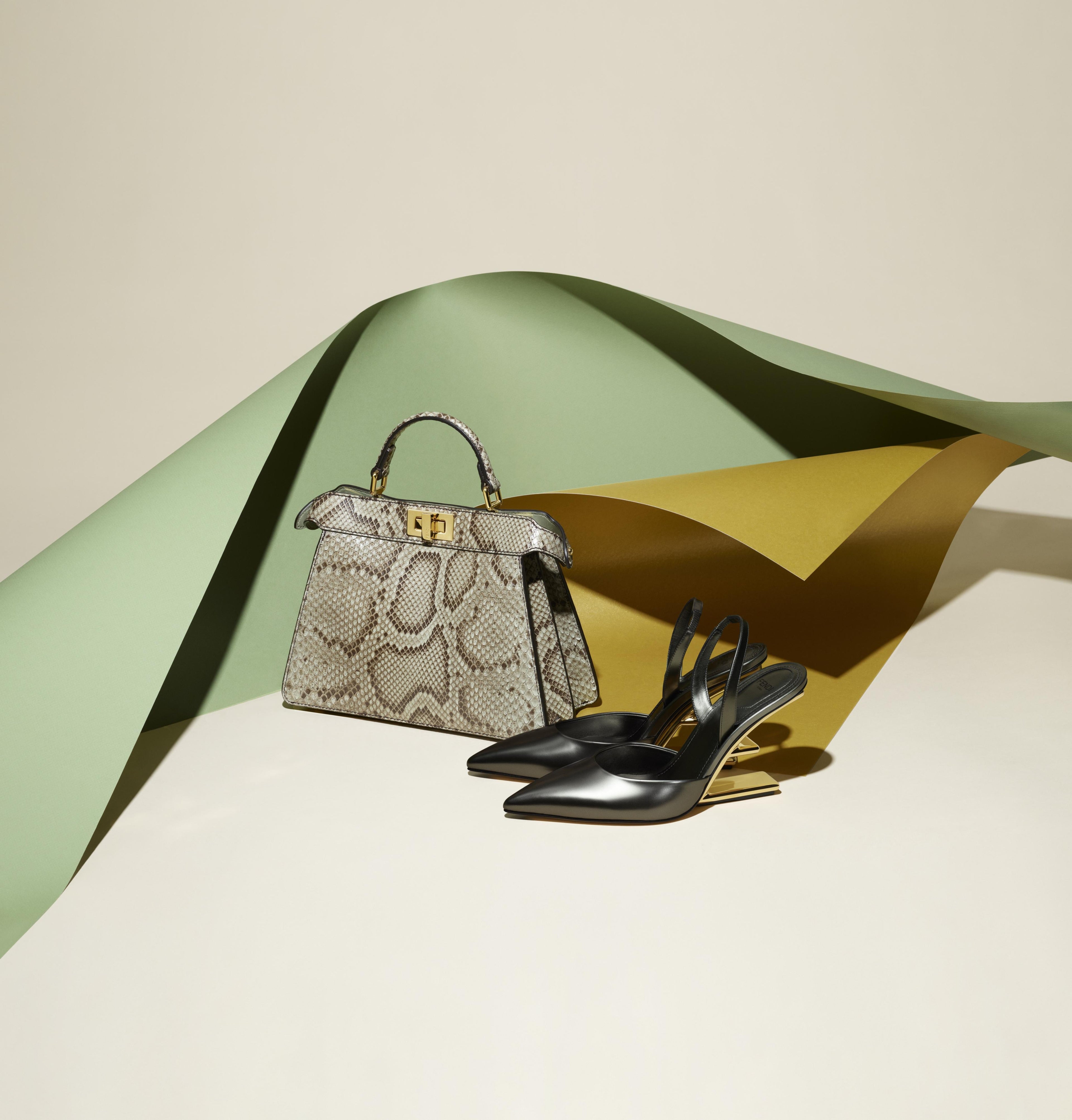 A structured snakeskin handbag with gold hardware sits beside a pair of black slingback high-heel pumps on a cream studio surface with sculptural green and gold paper drapes behind it.